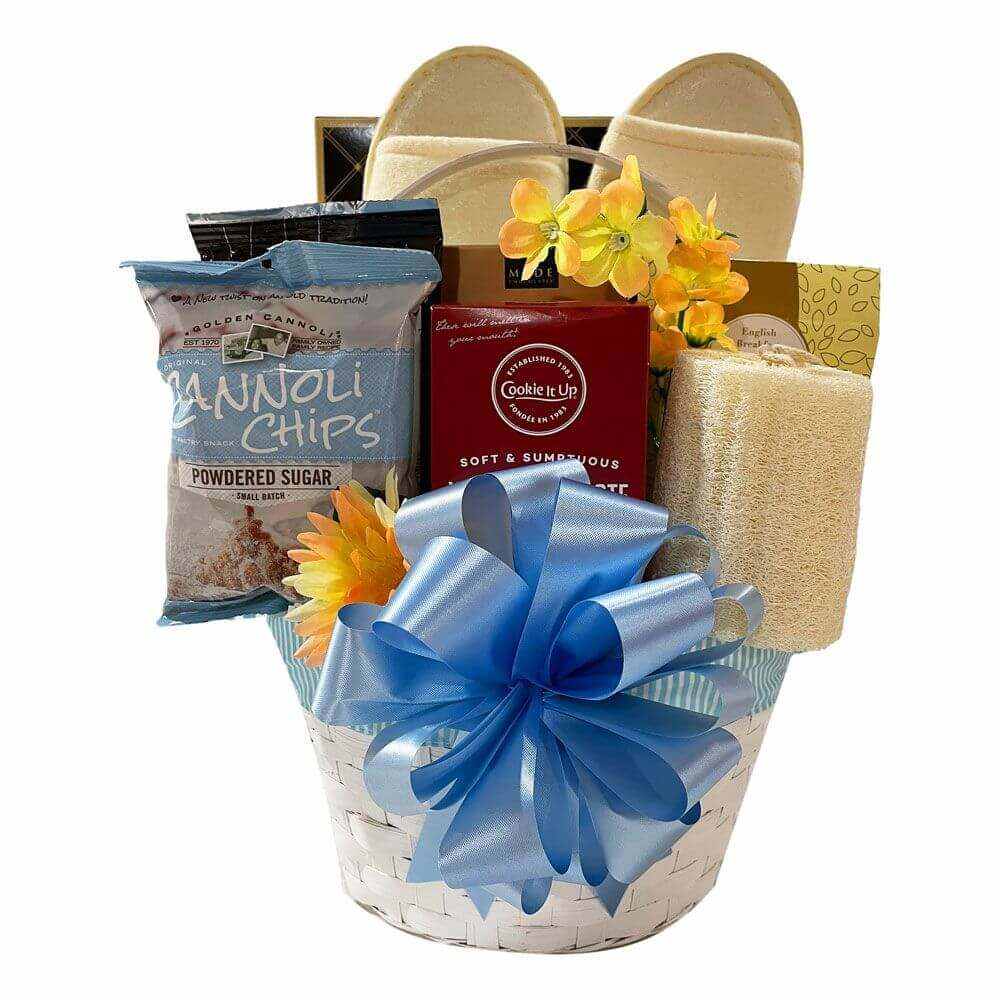 Pamper Gift Basket+ with bath slippers, loofah, cookies, chocolate, cannoli chips, brownies, lemon drops, tea, coffee, and shortbread in a decorative white basket with blue ribbon
